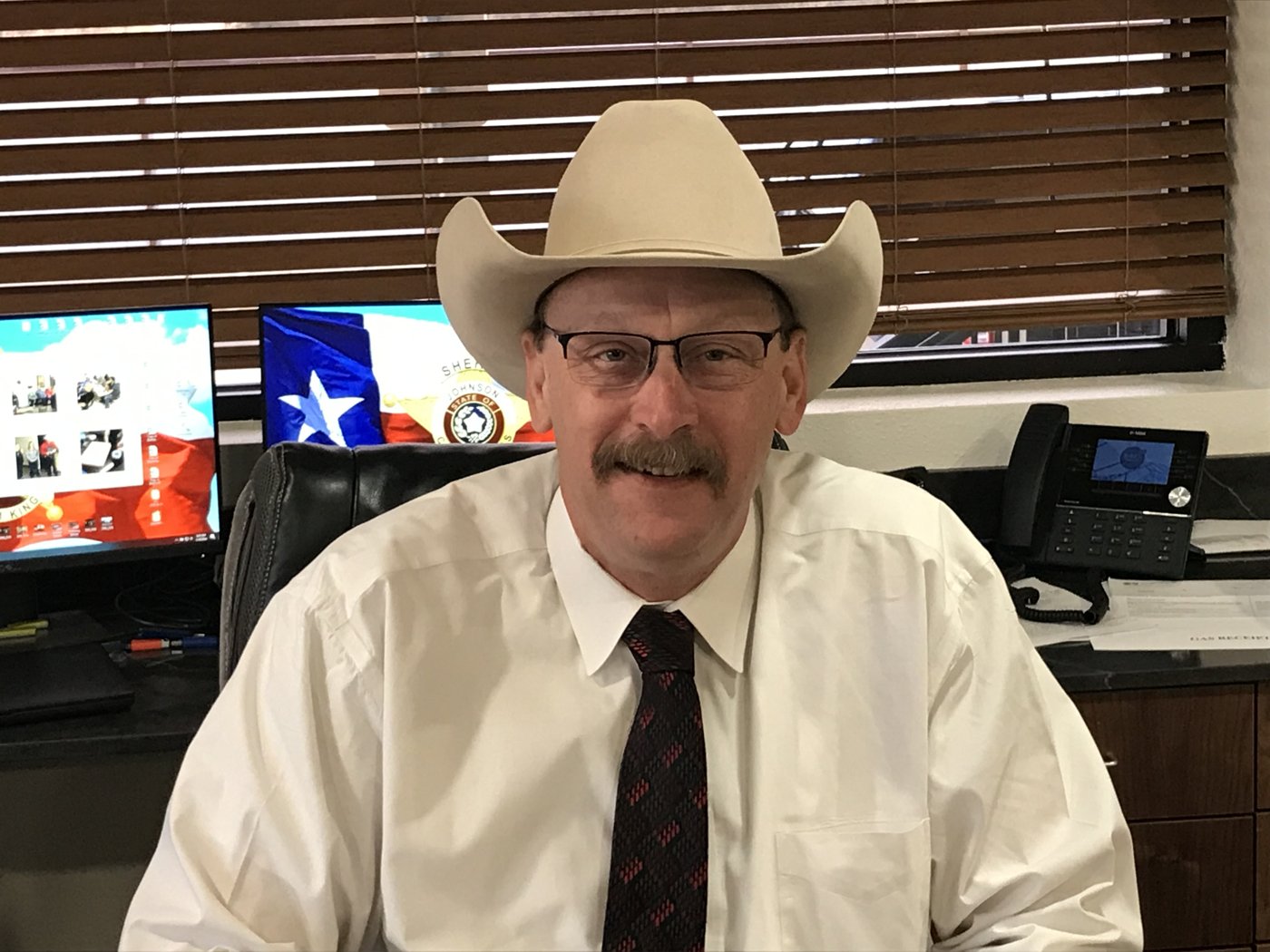 Sheriff Adam King at his desk at the Johnson County Sheriff&rsquo;s Office, with the Texas flag and Sheriff&rsquo;s badge behind him