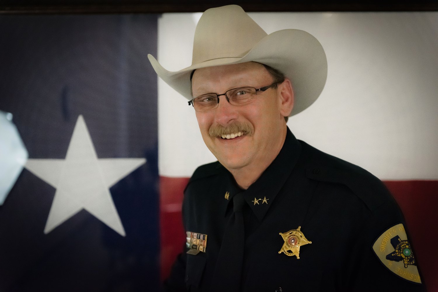 Sheriff Adam King of Johnson County, Texas, smiling in front of the Texas flag